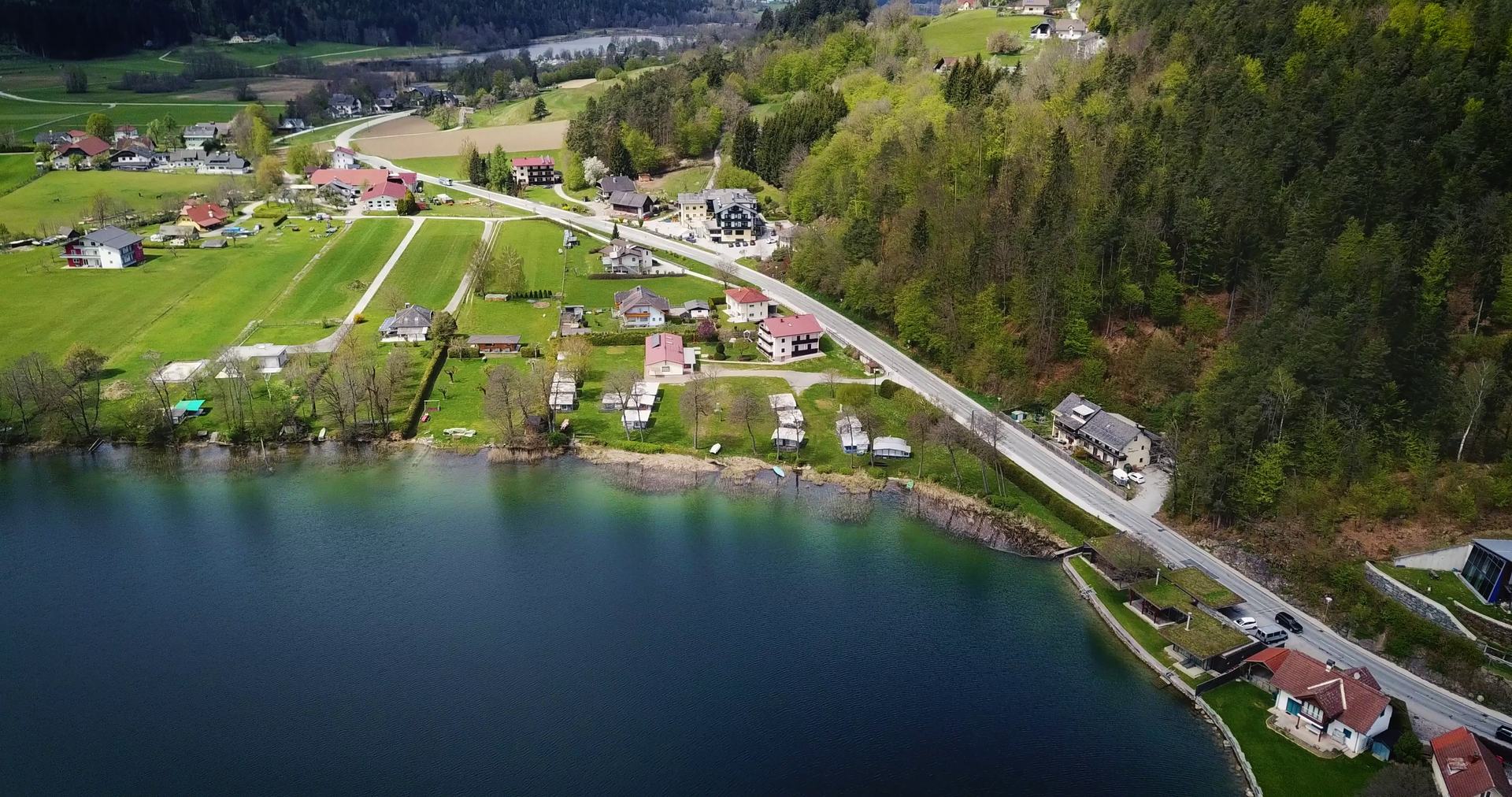 Drohnenaufnahme des Seeufers mit Uferstraße, Häusern und Wald